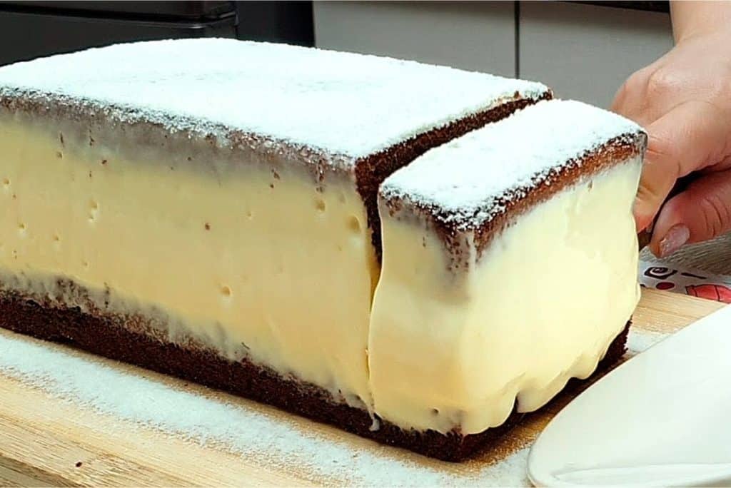 Bolo de leite retangular, coberto com açúcar de confeiteiro, sendo fatiado. A textura cremosa do recheio contrasta com a massa escura, criando uma sobremesa visualmente atraente.