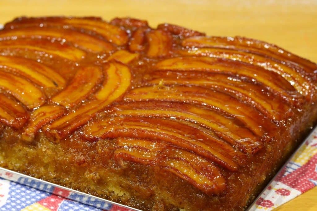 Bolo de banana caramelizada, servido em um prato retangular. O bolo tem uma camada de bananas caramelizadas no topo, criando uma aparência brilhante e apetitosa.