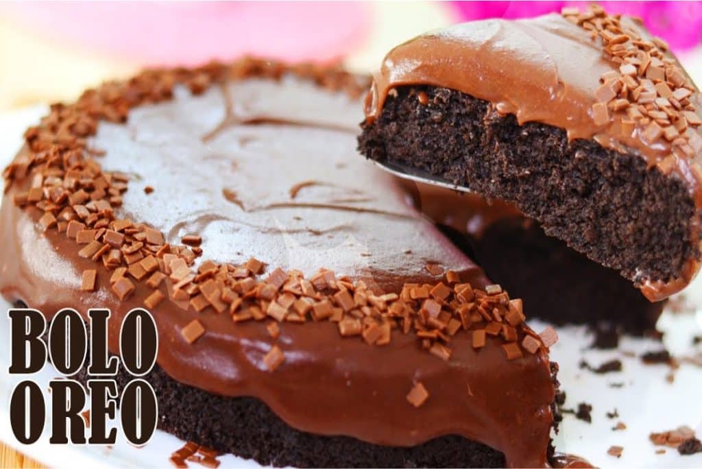 Bolo Oreo coberto com uma camada generosa de chocolate, decorado com granulado nas bordas. Uma fatia está sendo servida, destacando a textura úmida e o recheio rico de chocolate.