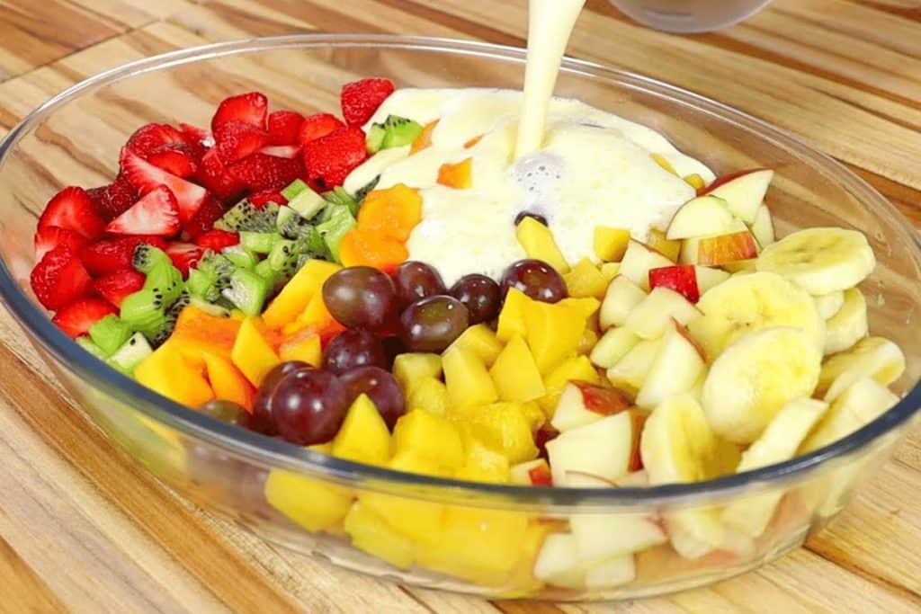 Salada de frutas cremosa em uma travessa de vidro, com uma variedade de frutas coloridas como morangos, kiwi, uvas, manga, banana e maçã, sendo cobertas com um creme branco.