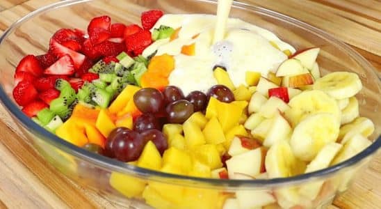 Salada de frutas cremosa em uma travessa de vidro, com uma variedade de frutas coloridas como morangos, kiwi, uvas, manga, banana e maçã, sendo cobertas com um creme branco.