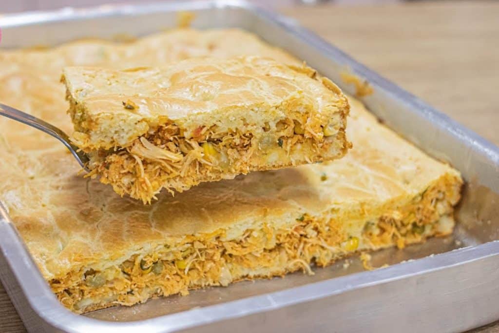 Torta de liquidificador, assada em uma forma retangular. Uma fatia está sendo retirada, mostrando o recheio suculento de frango desfiado.