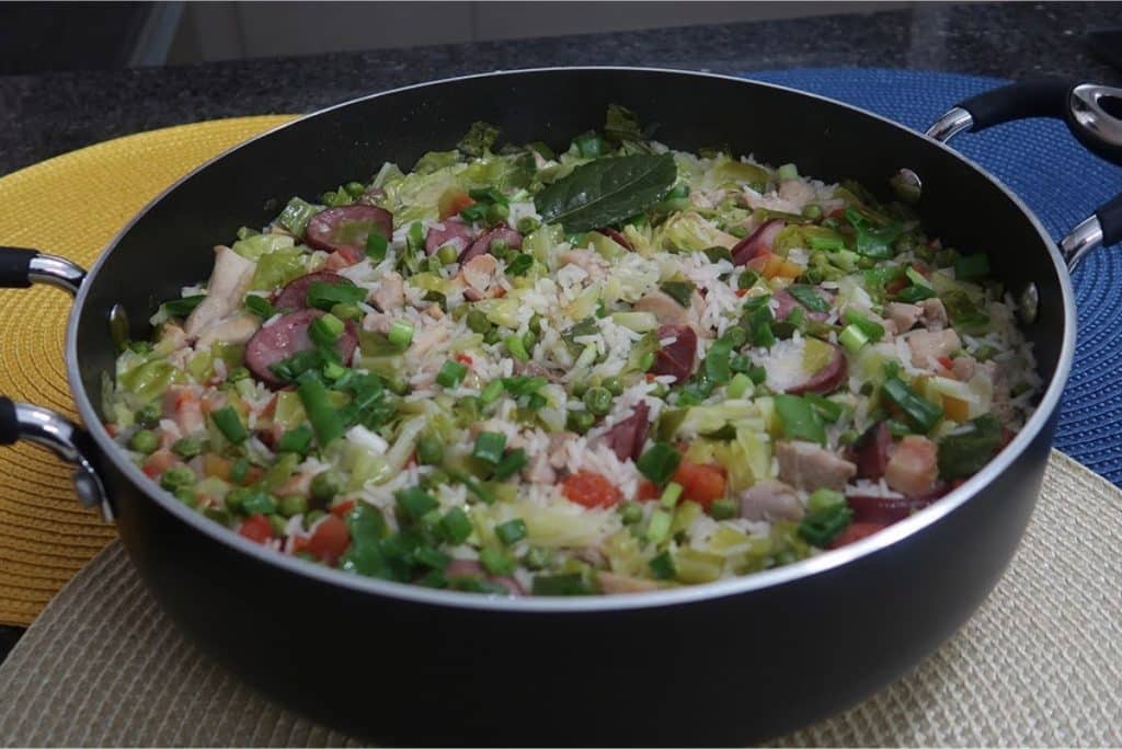 Uma panela grande com arroz à brasileira, cheia de legumes coloridos e pedaços de linguiça, decorada com ervas frescas.