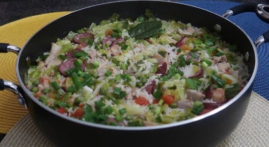 Uma panela grande com arroz à brasileira, cheia de legumes coloridos e pedaços de linguiça, decorada com ervas frescas.