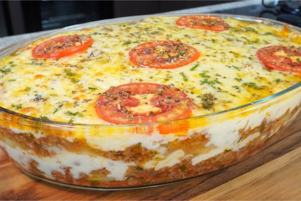 Uma travessa de vidro cheia de lasanha, com camadas de molho, carne, queijo e coberta com fatias de tomate e ervas frescas.