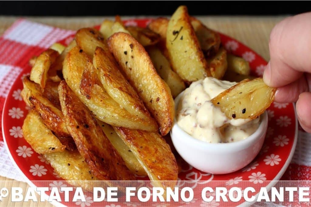 Batatas de forno crocantes, servidas em um prato vermelho com um molho cremoso em um pequeno recipiente ao lado. Uma mão está prestes a mergulhar uma batata no molho