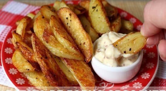 Batatas de forno crocantes, servidas em um prato vermelho com um molho cremoso em um pequeno recipiente ao lado. Uma mão está prestes a mergulhar uma batata no molho