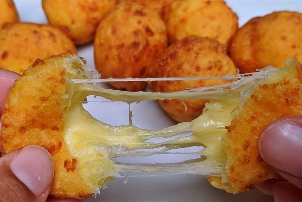 Bolinha de queijo com maisena, com queijo derretido se esticando ao ser aberta. Ao fundo, outras bolinhas douradas e crocantes, formando um delicioso aperitivo.