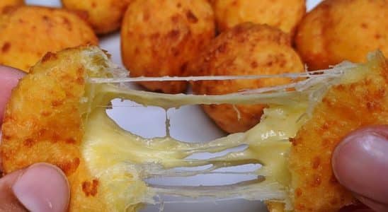 Bolinha de queijo com maisena, com queijo derretido se esticando ao ser aberta. Ao fundo, outras bolinhas douradas e crocantes, formando um delicioso aperitivo.