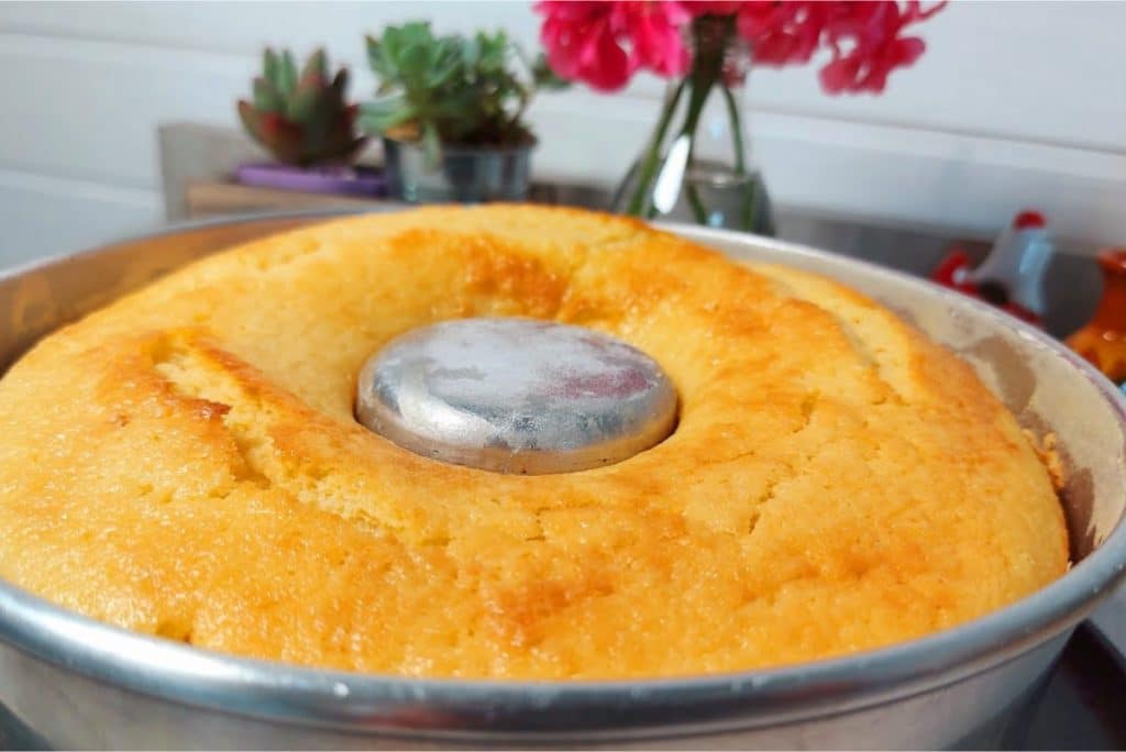 Bolo de limão dourado e fofinho, recém-saído do forno, em uma forma de furo central. Ao fundo, uma cozinha decorada com vasos de suculentas e flores vermelhas, criando um ambiente acolhedor.