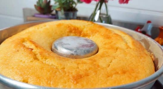 Bolo de limão dourado e fofinho, recém-saído do forno, em uma forma de furo central. Ao fundo, uma cozinha decorada com vasos de suculentas e flores vermelhas, criando um ambiente acolhedor.