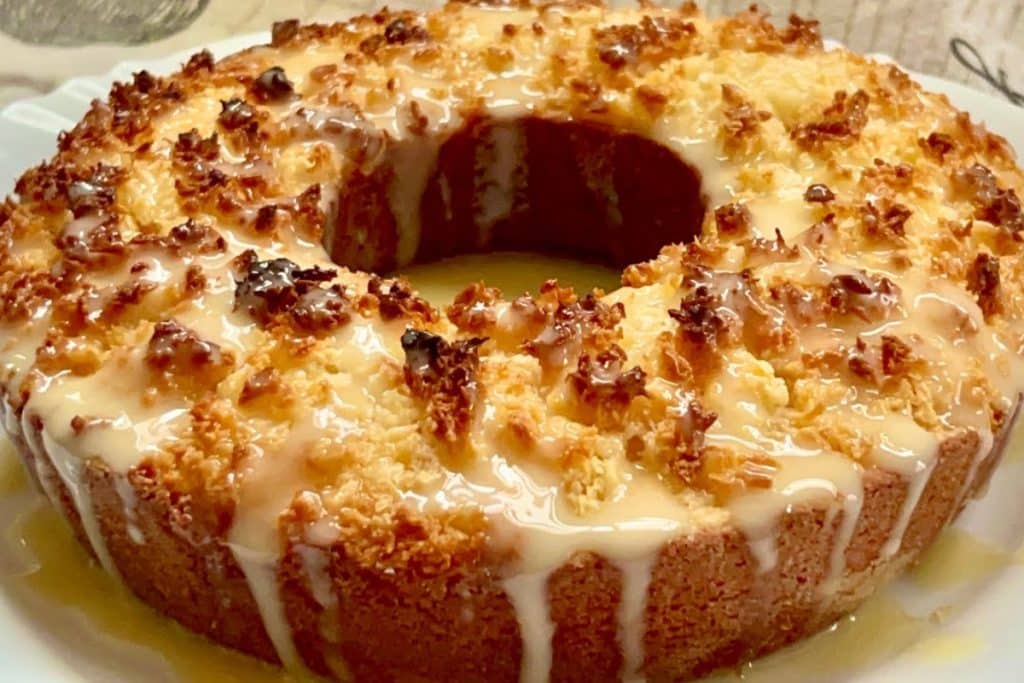 Bolo pão de coco, assado em forma de furo central, com cobertura de coco dourado e uma calda branca escorrendo pelas laterais. O bolo tem uma aparência úmida e deliciosa