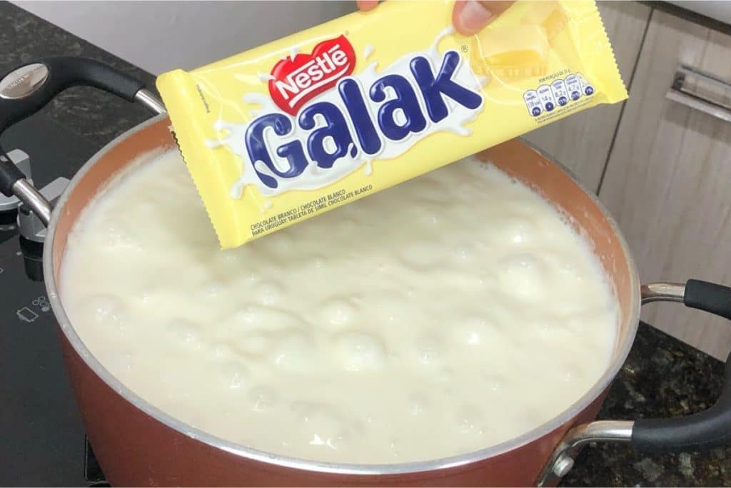 Canjica com chocolate branco sendo preparada em uma panela, com uma barra de chocolate branco Galak pronta para ser adicionada à mistura cremosa e quente.