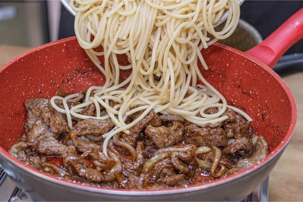 Macarrão ao bife acebolado, com espaguete sendo adicionado a uma frigideira vermelha cheia de bife suculento e cebolas caramelizadas, criando uma combinação saborosa.