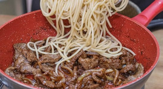 Macarrão ao bife acebolado, com espaguete sendo adicionado a uma frigideira vermelha cheia de bife suculento e cebolas caramelizadas, criando uma combinação saborosa.