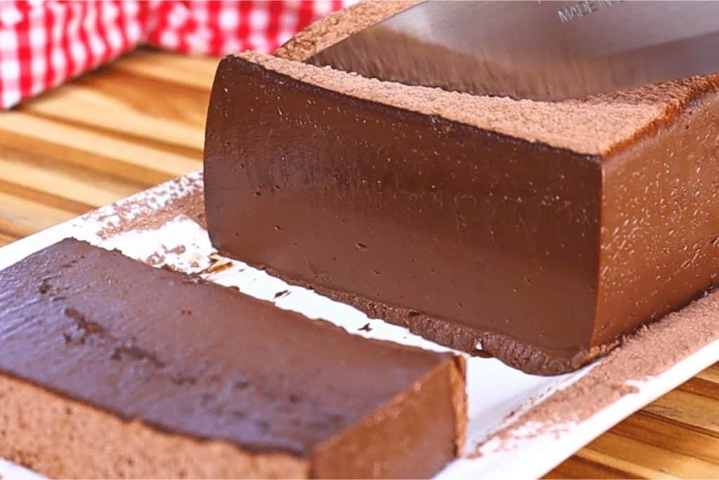 Mousse de chocolate de cortar, com uma textura firme e brilhante, servido em uma travessa. Uma fatia foi cortada, revelando a cremosidade e o rico sabor do chocolate. Ao fundo, um pano de prato xadrez vermelho e branco.