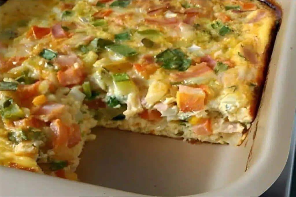 Omelete de forno colorido, com pedaços de legumes e temperos, servido em uma travessa retangular. Uma porção foi retirada, revelando sua textura fofa e suculenta, repleta de ingredientes frescos.