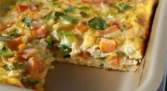 Omelete de forno colorido, com pedaços de legumes e temperos, servido em uma travessa retangular. Uma porção foi retirada, revelando sua textura fofa e suculenta, repleta de ingredientes frescos.