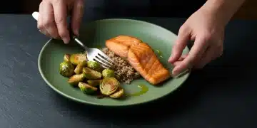 Close-up of a person's hands preparing a balanced meal with salmon, quinoa, and roasted vegetables on a dark green plate.