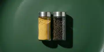 Overhead view of two minimalist spice jars, one with turmeric powder and one with whole black peppercorns, on a forest-green surface, illustrating ingredient synergy.